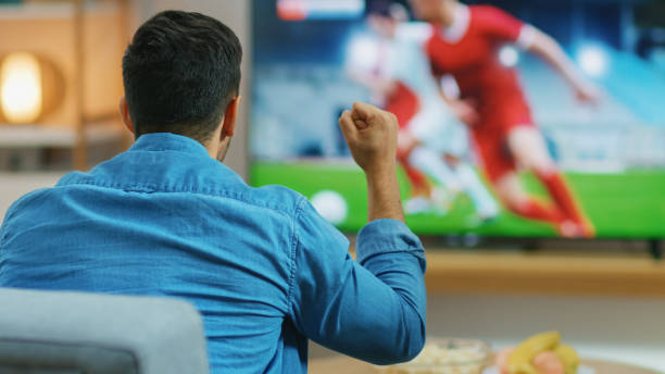 ホームスポーツファンはテレビで重要なサッカーの試合を見て、彼は積極的に彼のチームを応援し、拳を握ります。テーブルの上にスナックやドリンクを提供する居心地の良いリビングルー� - fan television football watching tv ストックフォトと画像
