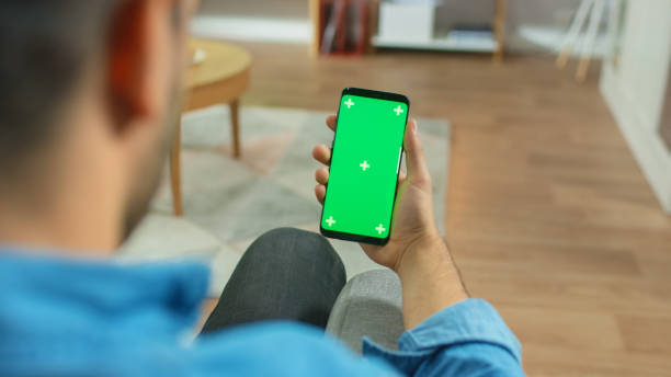 소파에 앉아 집에서 남자는 녹색 크로마 키 화면 터치 스크린 스마트 폰을 보유하고있다. 소셜 네트워크를 통해 검색, 뉴스를 읽거나 응용 프로그램을 사용합니다. - key house house key green 뉴스 사진 이미지