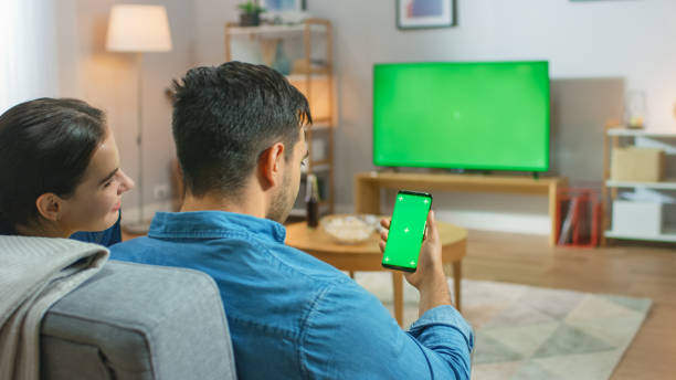 Happy Couple Sitting At Home in the Living Room Watching Green Chroma Key Screen Television, Relaxing on a Couch. Guy also Uses Green Mock-up Screen Smartphone. Happy Couple Sitting At Home in the Living Room Watching Green Chroma Key Screen Television, Relaxing on a Couch. Guy also Uses Green Mock-up Screen Smartphone. chroma key photos stock pictures, royalty-free photos & images