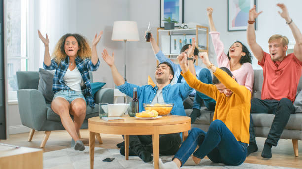 at home diverse group of sports fans sitting on couch watching important sports game match on tv, they cheer for team, celebrate victory after team scoring winning goal. cozy room with snacks drinks. - lanche da tarde imagens e fotografias de stock