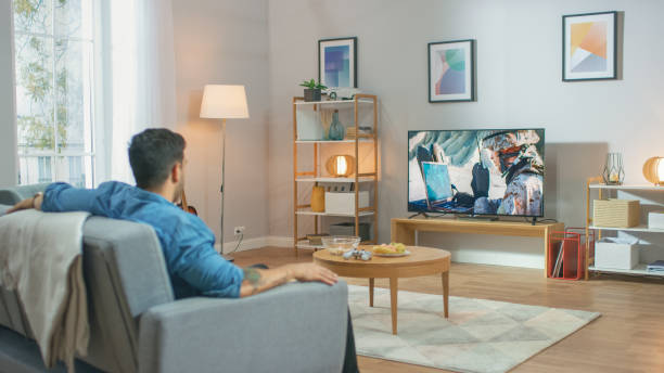 in the living room: guy relaxing on a couch watching war movie en un televisor. la acción de guerra militar moderna con el soldado de guerra se muestra en una televisión. - high definition television fotografías e imágenes de stock