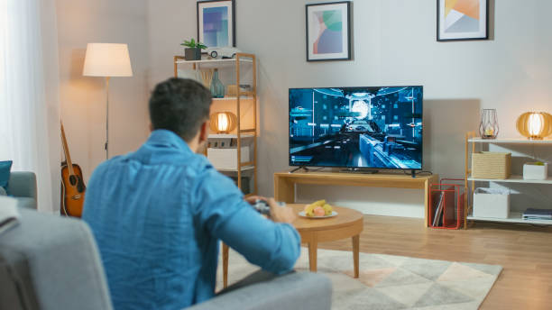 in the living room man sitting on a couch holds controller playing in a console video game, 3d action shooter gameplay shown auf tv-bildschirm. - multiplayer stock-fotos und bilder