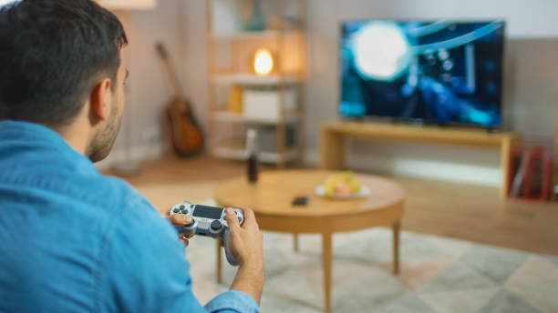 istock In the Living Room Man Sitting on a Couch Holds Controller Playing in a Console Video Game, 3D Action Shooter Gameplay Shown on TV Screen. 1178292492