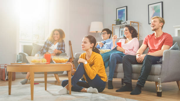 w domu diverse group friends oglądanie telewizji razem, jedzenie przekąski i picie napojów. prawdopodobnie oglądają grę sportową, film lub sitcom tv show. młodzi ludzie bawią się razem. - friends television show zdjęcia i obrazy z banku zdjęć