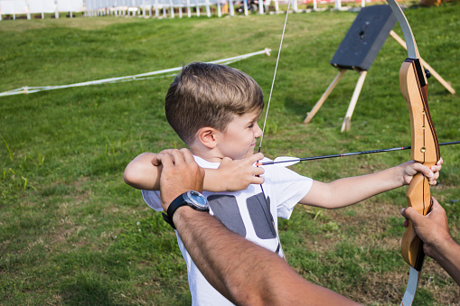Small kid with bow and arrow learning to aim at the target with help of archery instructor.