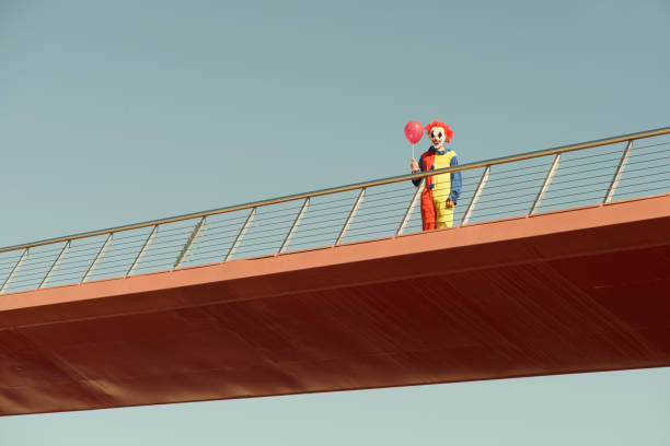 a creepy clown wearing a colorful yellow, red and blue costume, holding a red balloon in his hand, standing in a bridge outdoors