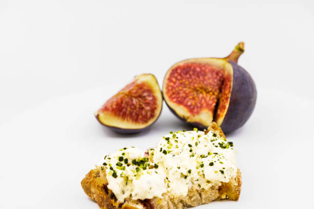 Fresh goat cheese tartine with garlic herbs and chopped shallots on a slice of cereal and dried fruit bread with a fresh fig - isolated on a white background stock photo