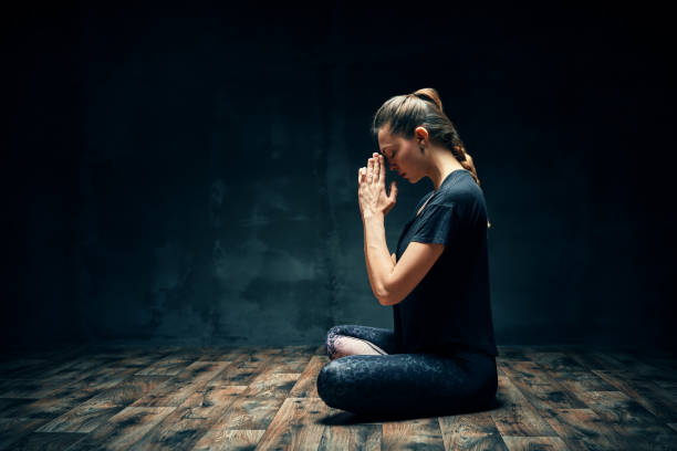 widok z tyłu młodej kobiety praktykującej jogę siedzącą w lotosie pozują z namaste w ciemnym pokoju i kopiuj przestrzeń - spirituality yoga zen like meditating zdjęcia i obrazy z banku zdjęć