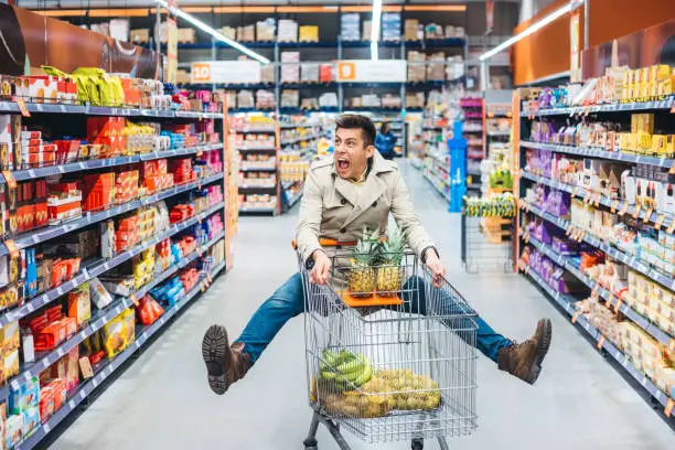 Young handsome Caucasian man jumping in grocery store and holding shopping cart.