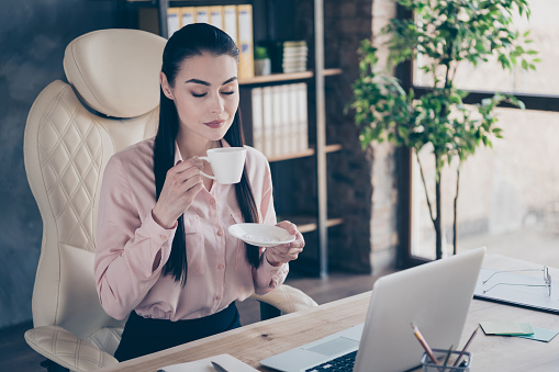 Photo of dreamy, cheerful resting marketer holding cup of warm beverage with her hands eyes closed with rejoicing at desktop with computer on it