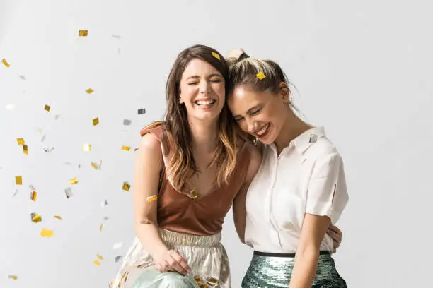 Portrait of two beautiful laughing young women hugging and celebrating under confetti.