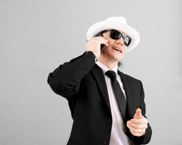 Successful attractive businessman in a suit, in a white hat and sunglasses, talking on a smartphone, smiles on a gray background. On the way to success. Business life. stock photo