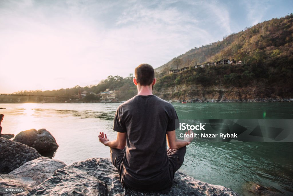 A young man meditates in a lotus position over the Ganges river in Rishikesh at the sunset. Meditating Stock Photo