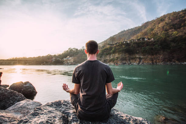 A young man meditates in a lotus position over the Ganges river in Rishikesh at the sunset. A young man meditates in a lotus position over the Ganges river in Rishikesh at the sunset. Meditation   stock pictures, royalty-free photos & images