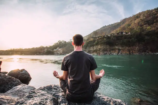 Photo of A young man meditates in a lotus position over the Ganges river in Rishikesh at the sunset.