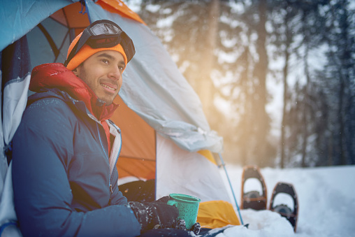Millenial indian guy setting camp with a tent in a snow filles canadian park during a snowshoeing trip