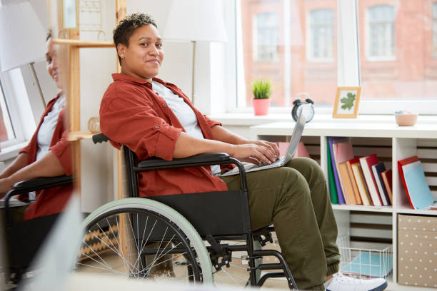 Disabled woman communicating online Portrait of African young woman with disabilities sitting in wheelchair and looking at camera while using laptop computer at home wheelchair stock pictures, royalty-free photos & images