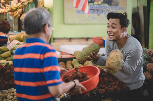 an asian male fruit stall owner selling jack-fruit to a chinese senior man at his stall