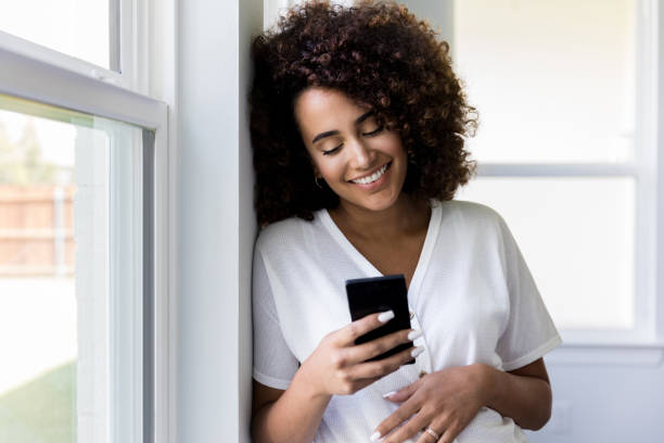 standing at window. pregnant woman smiles and checks phone As she stands by the windows of her new house, the mid adult pregnant women smiles as she checks her smart phone. scrolling photos stock pictures, royalty-free photos & images