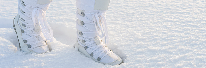 Female legs with white snow boots on, walking by the snow cover in sunlight. Winter panorama background.