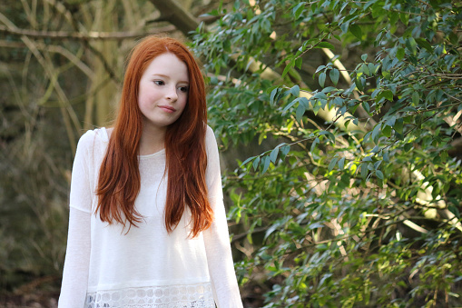 Stock photo showing a beautiful, ginger haired teenager wearing denim shorts and lacy white top, looking at water flowing under a wooden bridge. Hands resting on wooden rail, looking down into water.
