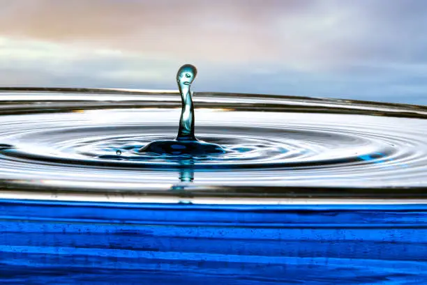 Single drop of water rises out of a calm pool surface into a column appearing to have two faces that show anger.