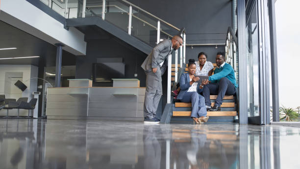 vier professionelle männliche und weibliche mitarbeiter sitzen auf der treppe lächelnd üben teamarbeit auf corporate tablet - africa business aboriginal computer stock-fotos und bilder