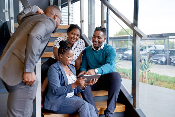 Four professional male and female coworkers sitting on staircase smiling practicing teamwork on corporate tablet stock photo