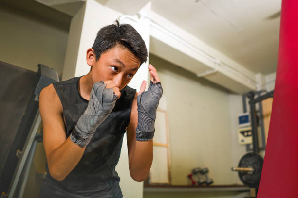 adolescent américain asiatique de boxe sur la gymnastique. jeune beau et féroce garçon de 13 ou 14 ans dans des enveloppements de poignet faisant la lutte d'entraînement poinçonnage sac lourd semblant frais et badass au club de forme physique - 13 14 years teenager 14 15 years child photos et images de collection