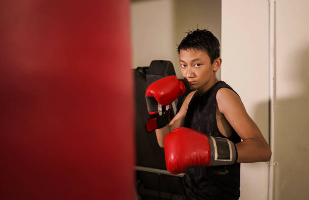 duro y fresco joven golpeando en la bolsa pesada. 13 o 14 años de edad adolescente asiático entrenamiento de boxeo tailandés buscando desafiante como un luchador malo practicando deporte en el club de fitness - 13 14 years fotografías e imágenes de stock
