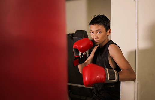 tough and cool young boy punching on heavy bag . 13 or 14 years old Asian teenager training Thai boxing workout looking defiant as a badass fighter practicing sport at fitness club