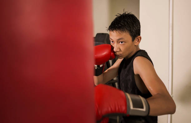 duro y fresco joven golpeando en la bolsa pesada. 13 o 14 años de edad adolescente asiático entrenamiento de boxeo tailandés buscando desafiante como un luchador malo practicando deporte en el club de fitness - 13 14 years teenager 14 15 years child fotografías e imágenes de stock