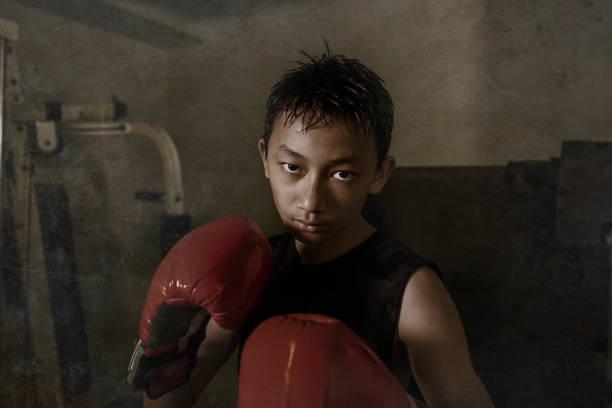 menino duro e fresco que perfura no saco pesado. 13 ou 14 anos de idade asiático adolescente treinamento tailandês boxe treino olhando desafiante como um lutador badass praticando esporte no clube de fitness - 13 14 years teenager 14 15 years child - fotografias e filmes do acervo