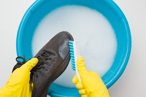 Hands in rubber protective gloves holding black sport shoe and brush above blue basin. Regular care about sneakers. Washing concept. Point of view shot. Closeup. Top view.