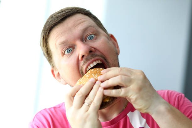 Funny bearded man with idiot facial expression eating big burger with enjoyment portrait