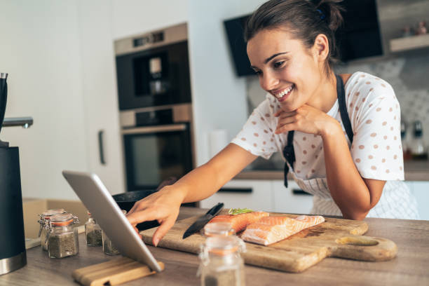 Online recipe Young beautiful woman using digital tablet while cooking salmon filet in kitchen salmon seafood stock pictures, royalty-free photos & images