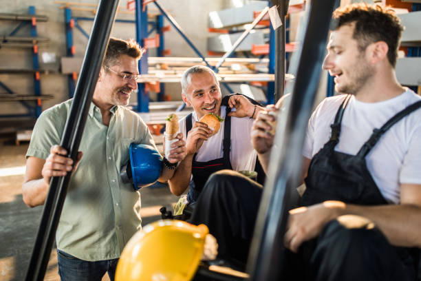 Happy manual workers and their manager talking on a lunch break in a factory. Group of workers communicating while eating sandwiches on a lunch break in industrial building. construction lunch break stock pictures, royalty-free photos & images