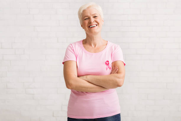 senior frau in rosa brustkrebs t-shirt stehend über weißer wand - breast cancer breast cancer awareness ribbon social awareness symbol human hand stock-fotos und bilder