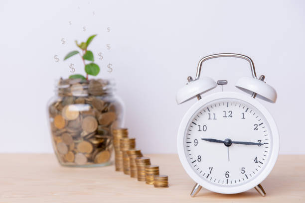 istock Savings on white background. Concept with white clock, jar and stack of money. This shows that savings are accumulating gradually and slowly. All on a wooden table and a white background. Copy space 1178522426