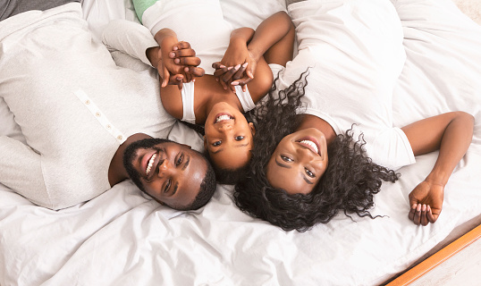 Happy family portrait. Young parents and their daughter lying in bed together, holding hands and looking to camera, top view