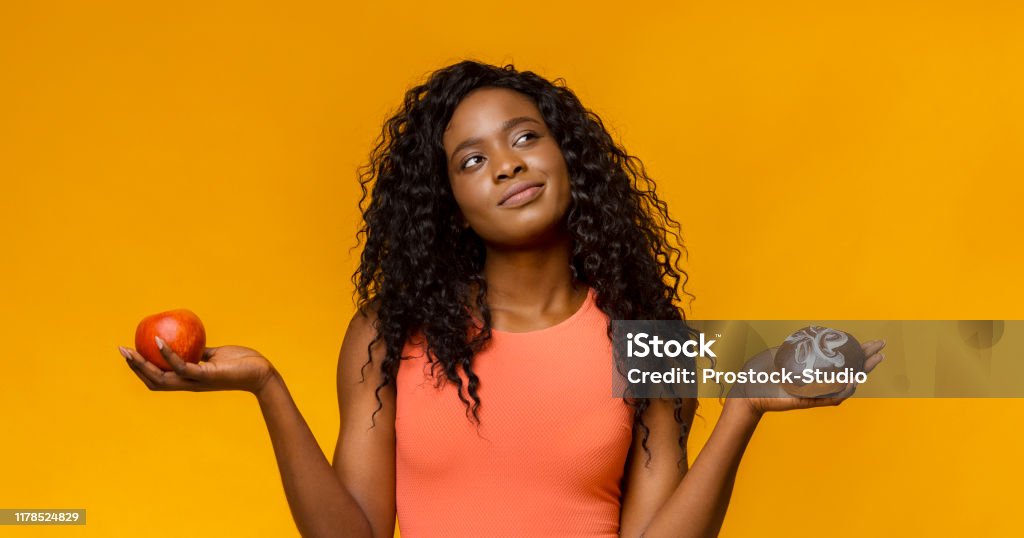 Young woman making choice, donut or apple Healthy lifestyle or nutrition concept with young african woman holding in hands apple and donut, yellow background, panorama Healthy Eating Stock Photo