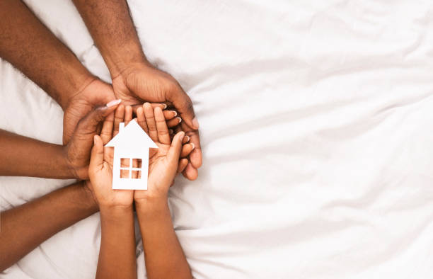 Closeup of paper house in hands of african american family Adoption and family housing concept. Cutout paper house in hands of african american family over white bed sheet background. Closeup, panorama adoption photos stock pictures, royalty-free photos & images