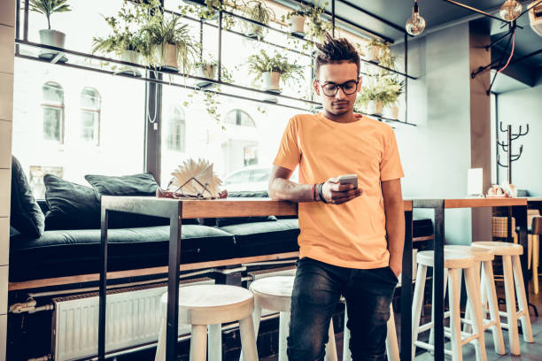 Young male indian freelancer in fast food cafe, handsome man reading text message during work on mobile phone in comfortable coffee shop. Panoramic windows background.