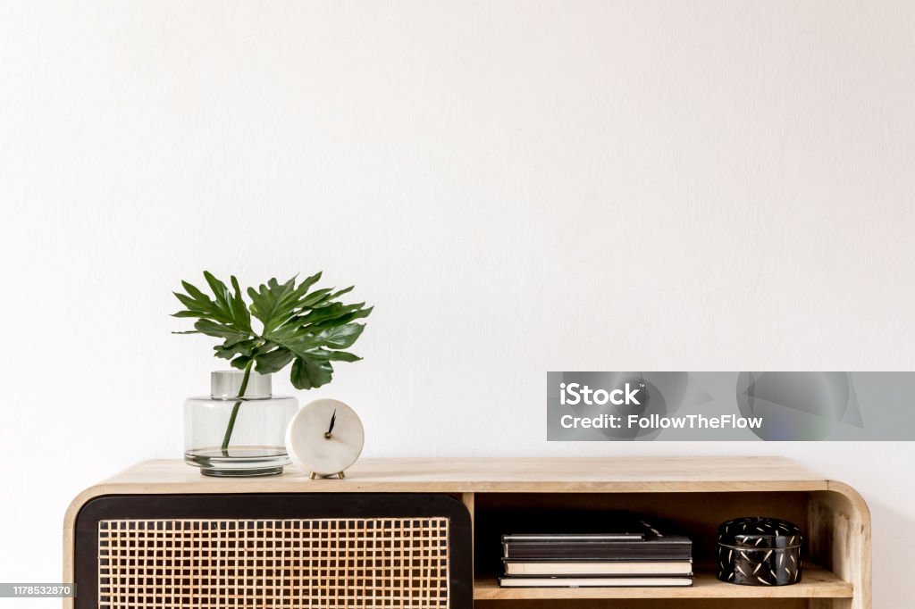 Interior design of living room at scandinavian apartment with stylish commode, tropical leaf in vase, books, white clock and elegant accessories. Modern home decor. Template. Copy space. White walls. Interior design of living room at nice scandinavian apartment with stylish furnitures and elegant accessories. Modern home decor. Template. Shelf Stock Photo
