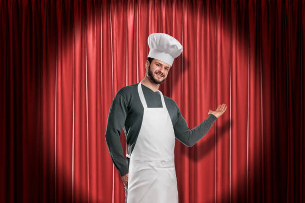 Young smiling handsome chef in white hat and apron lit up by limelight making presenting gesture at red stage curtain.
