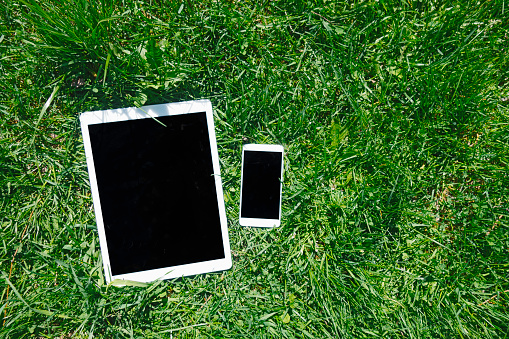 White smartphone and digital tablet on green grass. Mock up of gadgets with black displays.