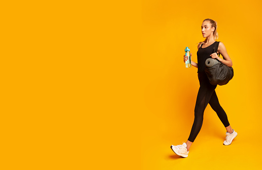 Woman Going To Gym Carrying Fitness Bag And Water Bottle On Yellow Studio Background. Full Length, Copy Space