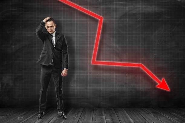 istock Businessman in black suit standing with one hand on head and with troubled expression on face at black graph ruled wall with red arrow pointing down. 1178543356
