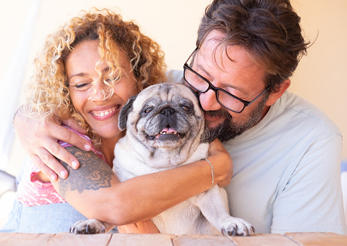 Beautiful couple man and woman in love with their clear pug dog. Forever friends. United and happy family. Smiling. Wooden table on terrace field