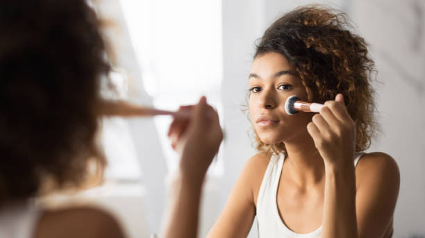 Afro Woman Applying Face Powder With Makeup Brush In Bathroom Makeup. Afro Young Woman Applying Face Powder With Cosmetic Brush Looking In Bathroom Mirror. Panorama, Selective Focus, foundation make up stock pictures, royalty-free photos & images